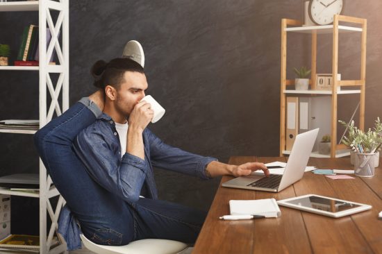 Short break for yoga in office. Flexible man practicing yoga at workplace, while typing on laptop and having coffee, copy space. Active employee at work, healthy lifestyle concept