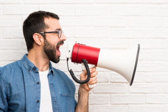 Handsome man with beard over white brick wall shouting through a megaphone