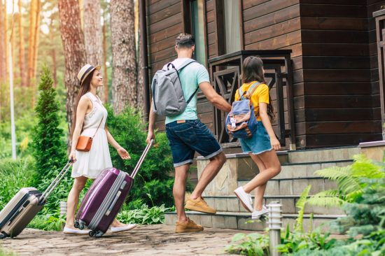 At last, hotel. A photo with sunny light effect of a glad family with suitcases and backpacs on a weekend coming to the hotel surrounded by trees, father holding his daughter's hand.
