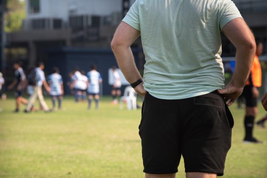 Father standing and watching his son playing football in a school tournament on a clear sky and sunny day. Sport, outdoor active, lifestyle, happy family and soccer mom and soccer dad concept.