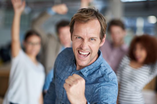 Hell yeah We did it. Mature man shouting triumphantly and making a fist with celebrating staff in the background