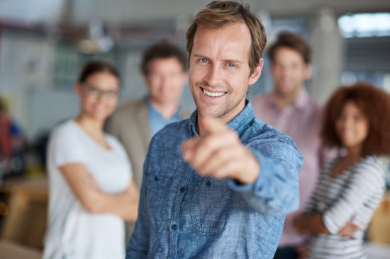 You could be our future colleague. Smiling man pointing at you a friendly group of office staff in the background